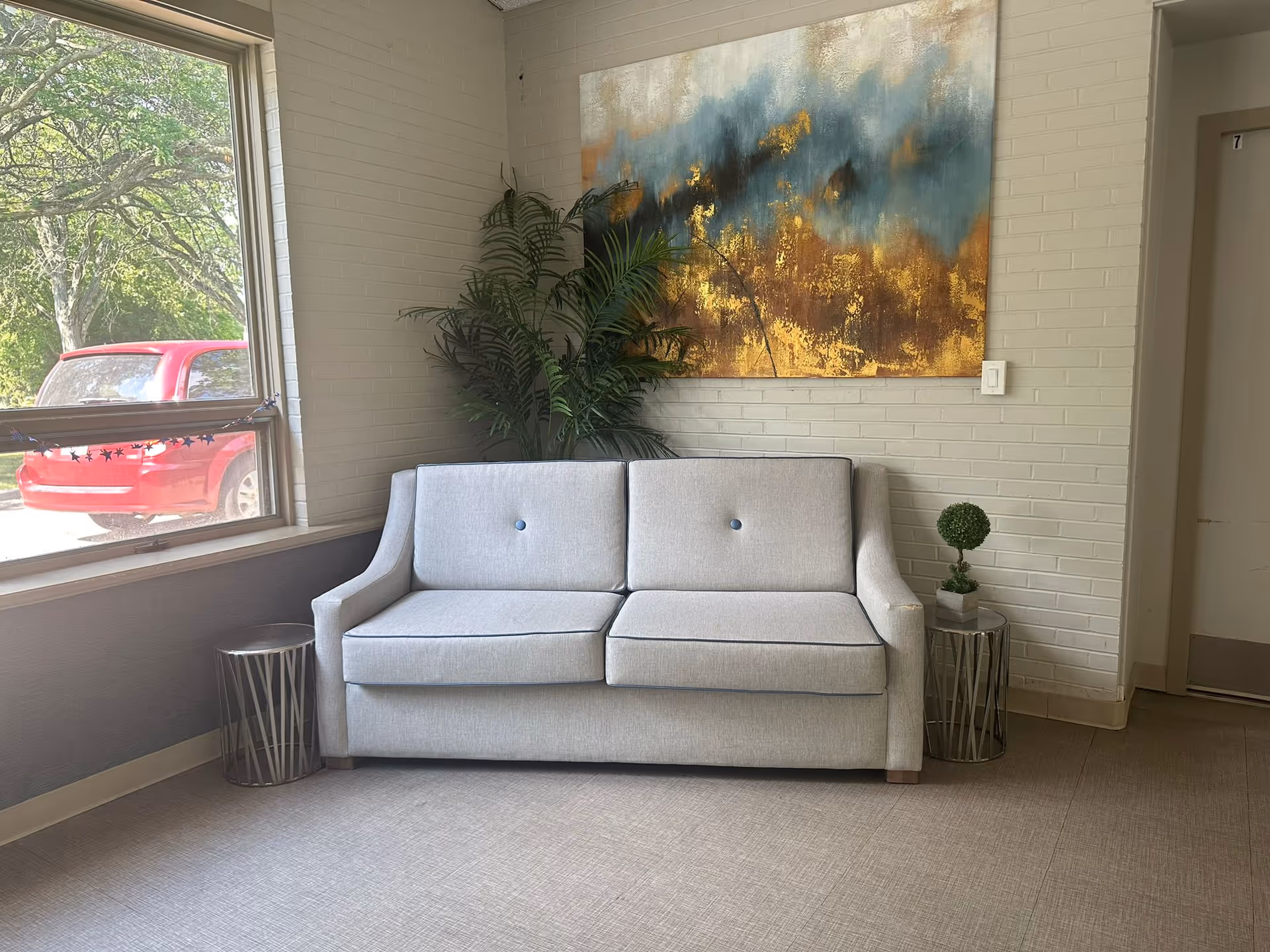 A small seating area with a light gray two-seater sofa placed against a white brick wall. Above the sofa hangs a large abstract painting with blue, gold, and brown tones. There is a green potted plant behind the sofa and a small round side table with a small topiary plant on the right side. A window on the left side shows a red vehicle and green trees outside. The floor is covered with light-colored tiles.