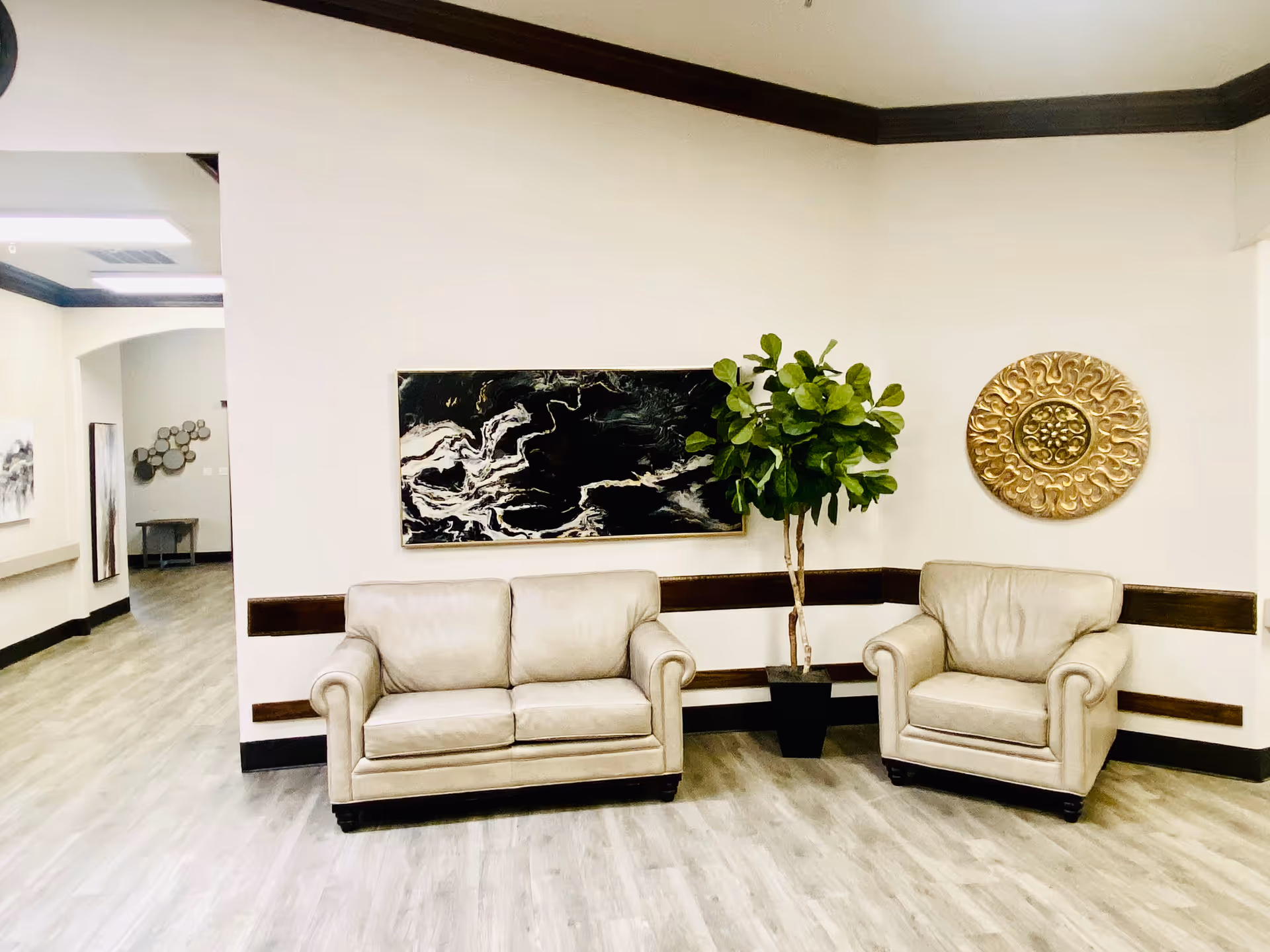 A seating area in a senior living facility with a beige loveseat and armchair, a potted green plant between them, a black and white abstract painting on the wall, and a decorative gold circular wall piece. The floor is light wood, and the walls are white with dark wood trim.