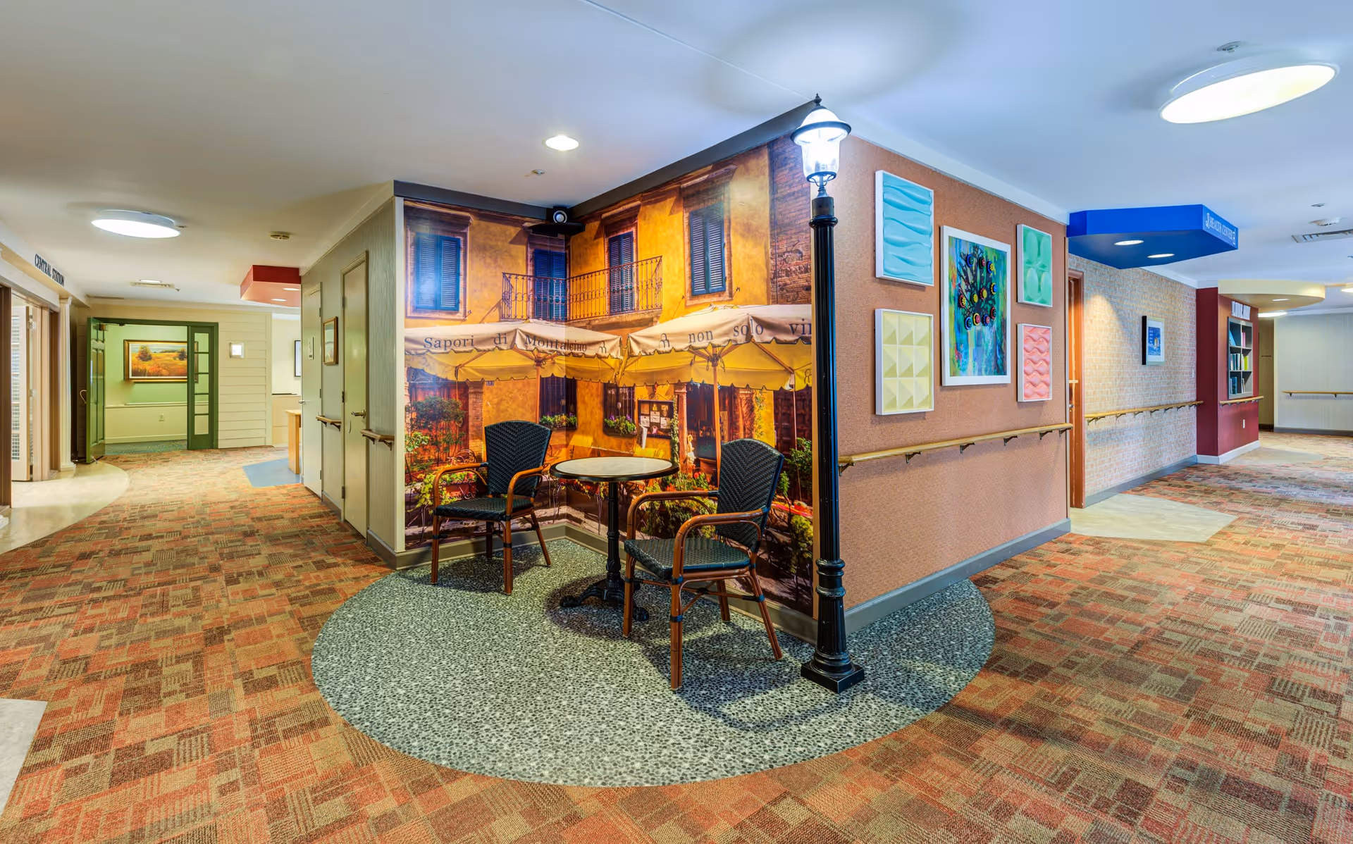 A hallway in a senior living facility with patterned carpet and handrails along the walls. There is a small seating area with two chairs and a round table in front of a wall mural depicting a European-style outdoor café scene. The seating area is accented by a decorative street lamp. The walls are decorated with colorful framed artwork. Doors and signage are visible along the hallway.