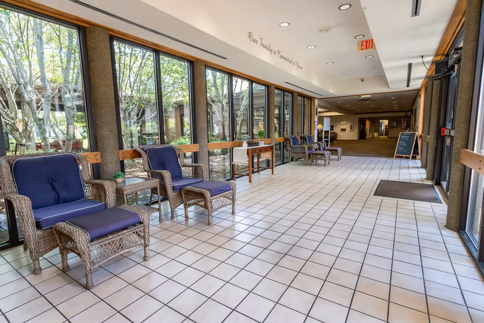A bright indoor seating area with large windows showing trees outside. The space has white tiled floors and several wicker chairs with blue cushions and matching footrests arranged along the windows. A small table with a plant and a welcome sign is also visible. The hallway extends into a carpeted area with additional seating and a chalkboard sign near the entrance door.