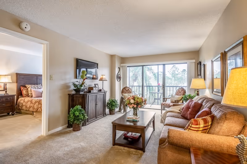 A cozy living room in a senior living facility with a brown sofa adorned with pillows, a wooden coffee table with a flower vase, two armchairs near a large window with a view of trees, a TV mounted on the wall above a wooden cabinet, and a glimpse into a bedroom with a bed and nightstand with a lamp.