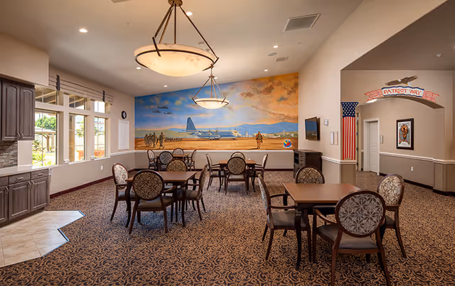A spacious dining area with several tables and chairs arranged on a patterned carpet. The room features a large mural on the far wall depicting a military scene with soldiers and an airplane. There are two hanging light fixtures from the ceiling, windows on the left side letting in natural light, and a sign above a hallway entrance that reads 'Patriot Way'.