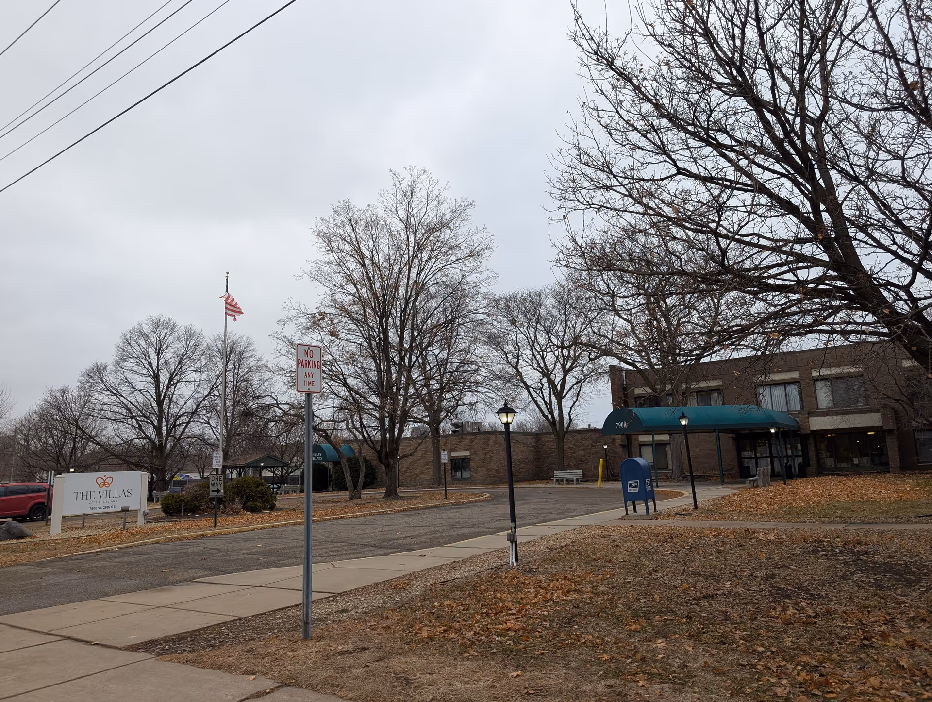 Exterior view of The Villas at The Cedars facility showing a brick building with a green awning over the entrance, a mailbox, a flagpole with an American flag, leafless trees, and a sign with the facility name and address.