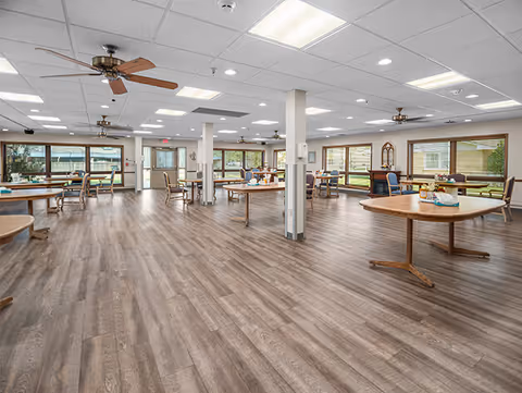 A spacious dining room with multiple round wooden tables and chairs arranged throughout. The room has large windows allowing natural light to fill the space, ceiling fans, and recessed lighting. The floor is covered with wood-patterned vinyl or laminate flooring.