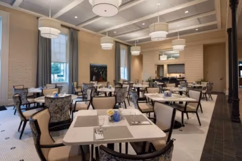 A spacious dining room with multiple tables and chairs arranged neatly. The tables are set with placemats, napkins, and small flower arrangements. Large windows with curtains allow natural light to fill the room. Modern pendant lights hang from the ceiling, and there is a serving area or counter at the back of the room.
