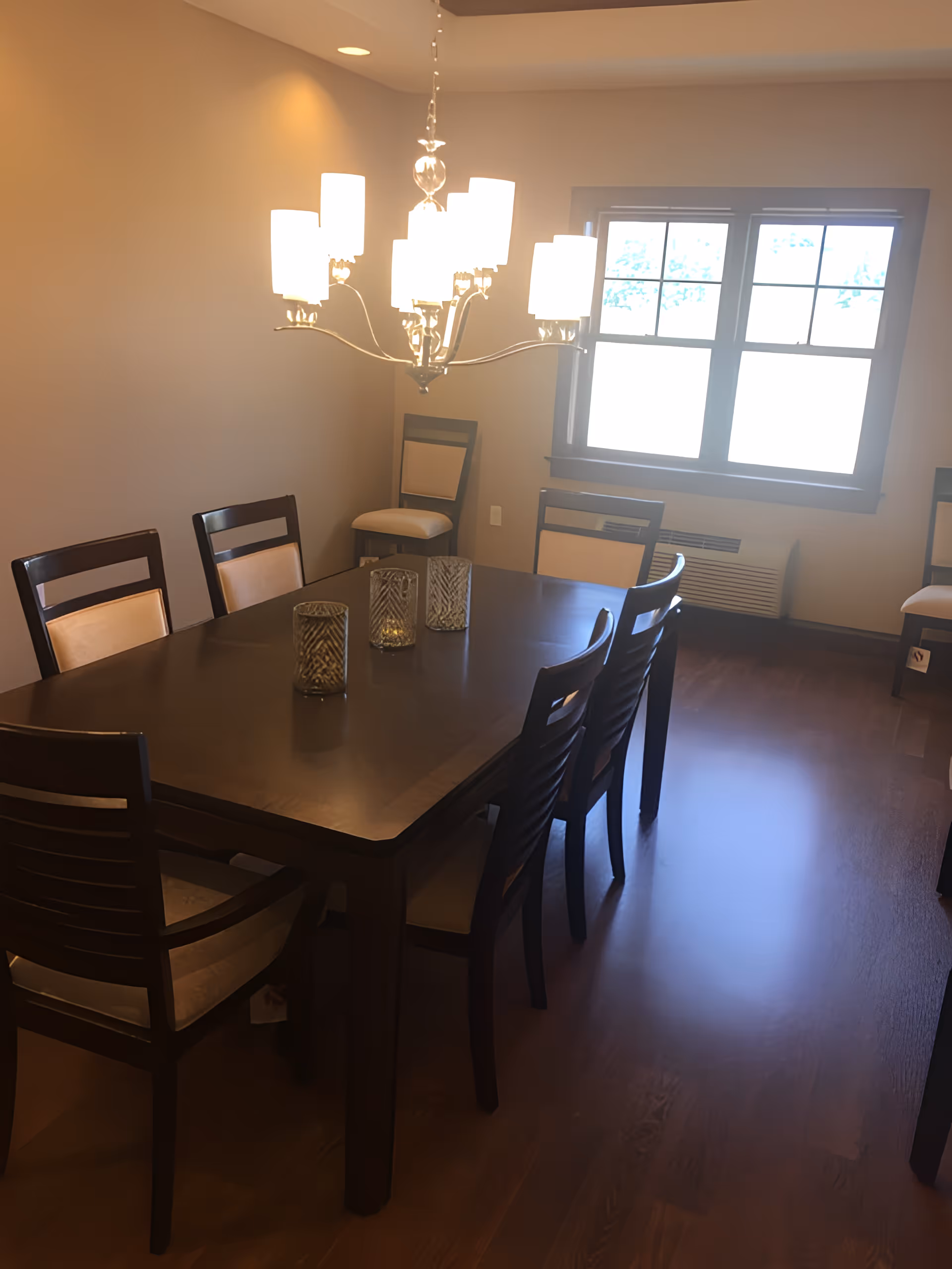 Interior dining room with a dark wooden table surrounded by chairs, a chandelier overhead and a window.
