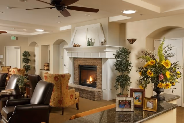 A cozy living room area with a lit fireplace surrounded by white mantel and brick. The room features comfortable armchairs, a ceiling fan, potted plants, and a granite countertop with framed photos and a vase of sunflowers. The walls are painted in neutral tones with arched doorways in the background.