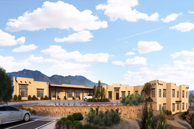 Exterior view of MorningStar Assisted Living & Memory Care of Santa Fe, a beige stucco building with southwestern architectural style, set against a backdrop of mountains and a partly cloudy blue sky. The building is surrounded by landscaped greenery and a parking area with cars.