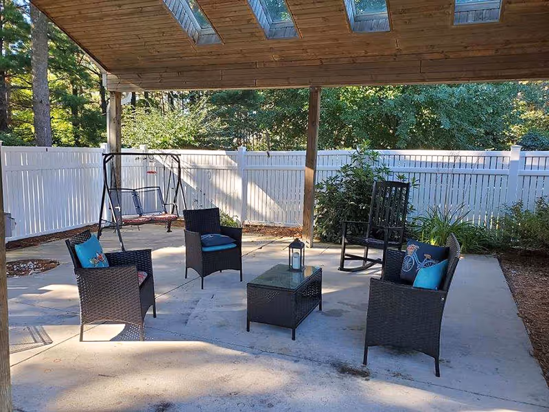 Covered outdoor patio with wicker chairs, a small table and a swing set against a white privacy fence and trees.