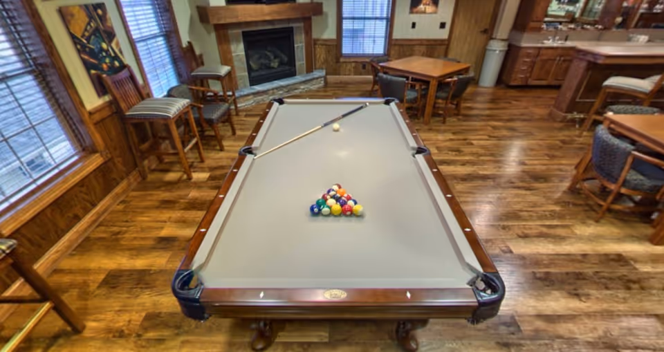 Pool table with racked balls and a cue in a wood-paneled common room with fireplace and seating.