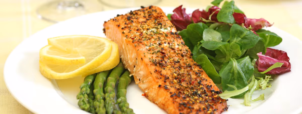 A white plate with a seasoned and cooked salmon fillet, several stalks of asparagus, two lemon slices, and a side of mixed leafy greens including spinach and radicchio.