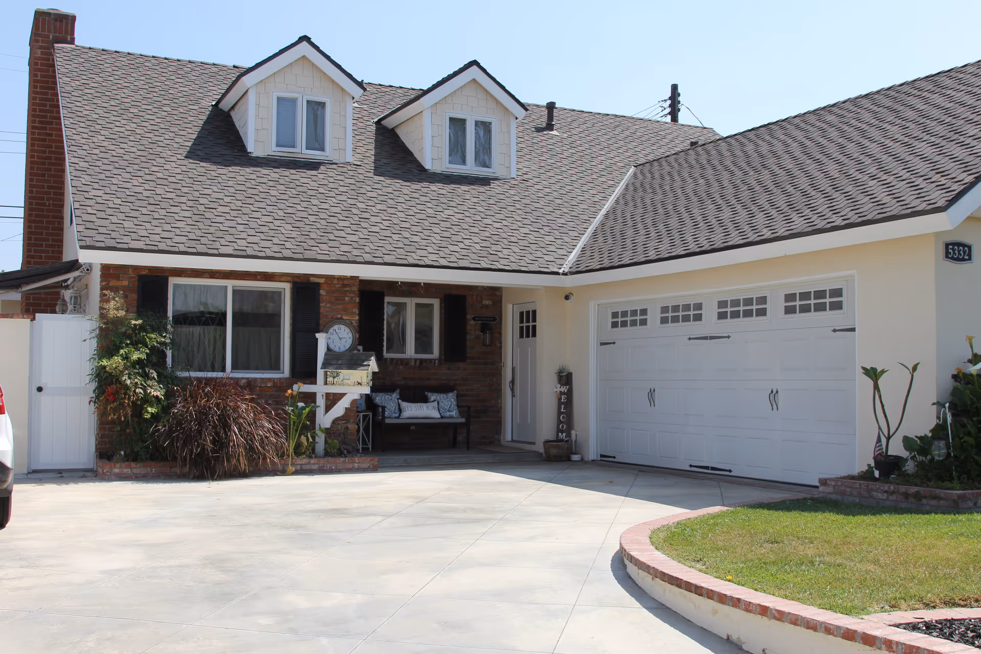 Front exterior view of a single-story house with a brick chimney, two dormer windows, a white garage door, a small porch with a bench, plants, and a concrete driveway.