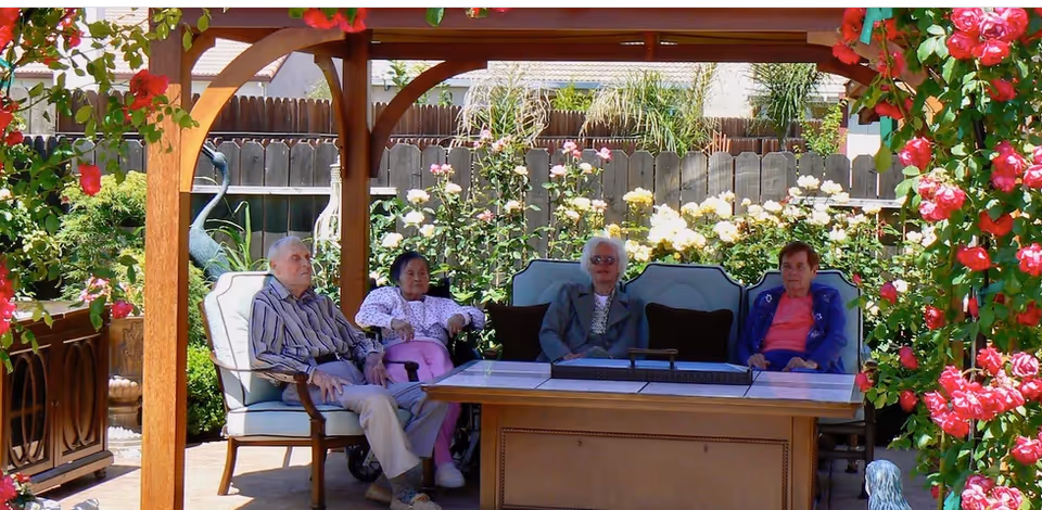Four elderly individuals sitting on cushioned outdoor chairs under a wooden pergola surrounded by blooming flowers and greenery in a garden setting.