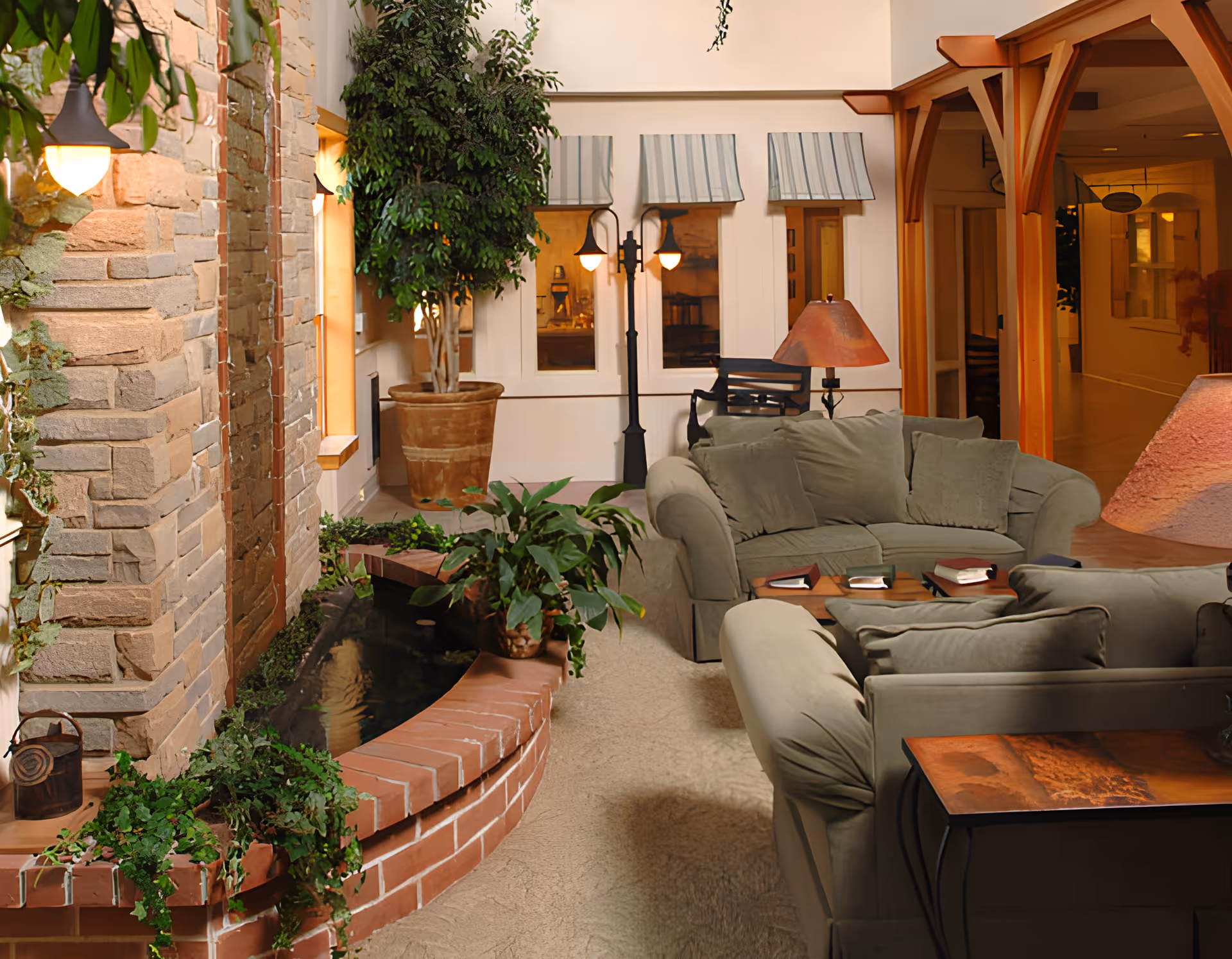 Indoor lounge area with beige sofas and cushions arranged around a wooden coffee table with books. There is a brick planter with green plants and a small water feature along a stone wall. Large potted plants and a lamp with a reddish shade add to the cozy atmosphere. The background shows windows with striped awnings and wooden archways leading to a hallway.