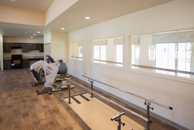 Interior view of a rehabilitation area with exercise equipment including stationary bikes and parallel bars for walking practice, adjacent to a kitchen area with dark cabinets and appliances. The room has large windows on one wall and wood-patterned flooring.