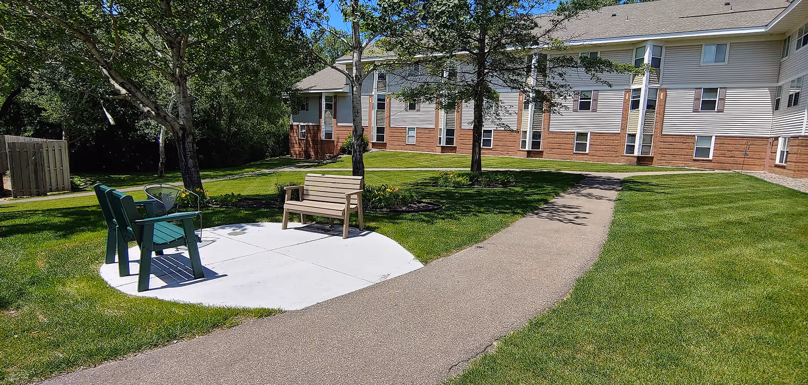 Outdoor seating area in a senior living facility with two green chairs and a wooden bench on a concrete pad surrounded by grass, trees, and a paved walkway leading to a multi-story building with white siding and brick accents.