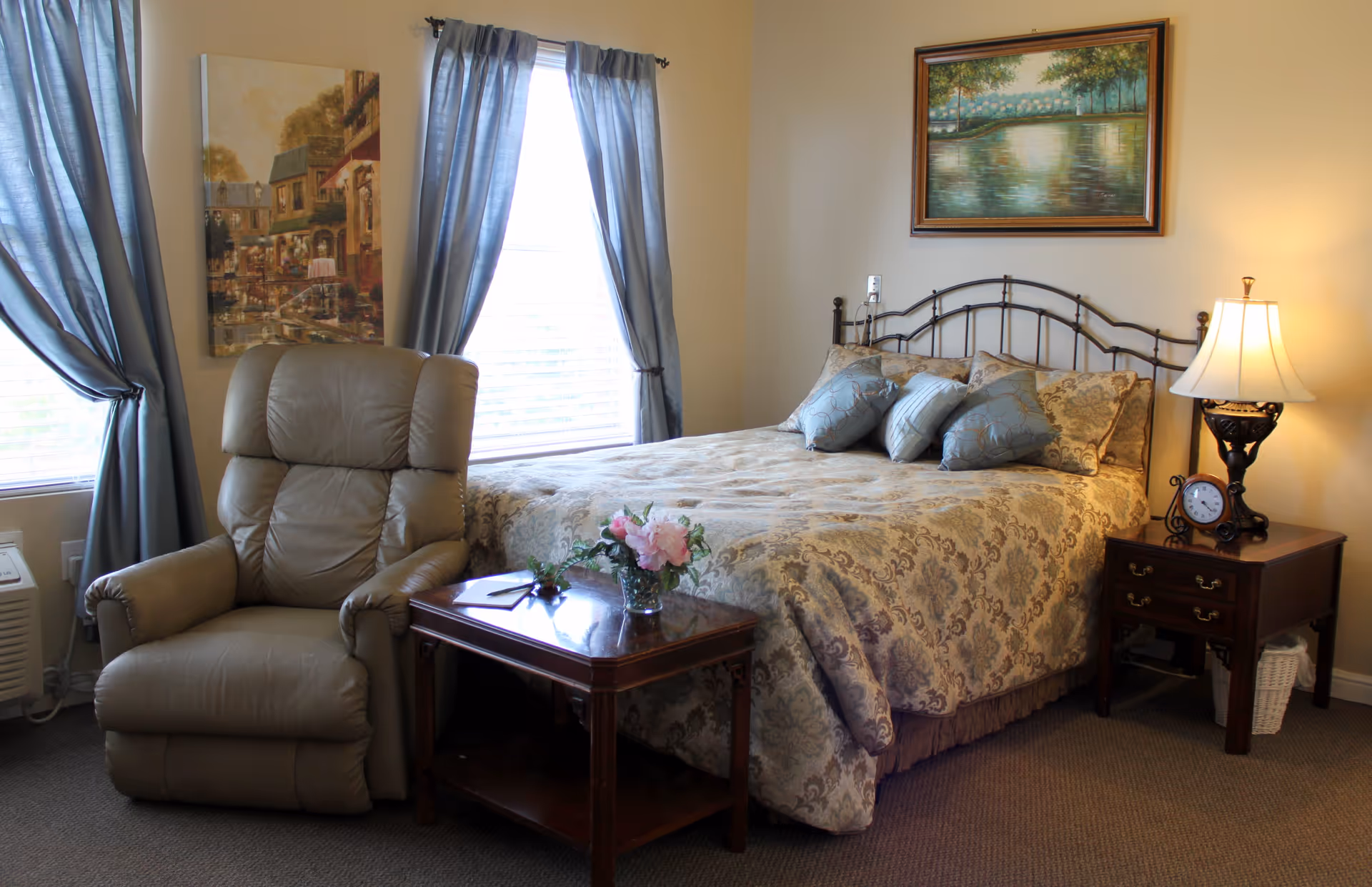 A cozy bedroom in Oakview Assisted Living & Memory Care featuring a large bed with patterned bedding and multiple pillows, a beige recliner chair, a wooden side table with a flower vase, a nightstand with a lamp and a clock, two windows with blue curtains, and two framed paintings on the walls.