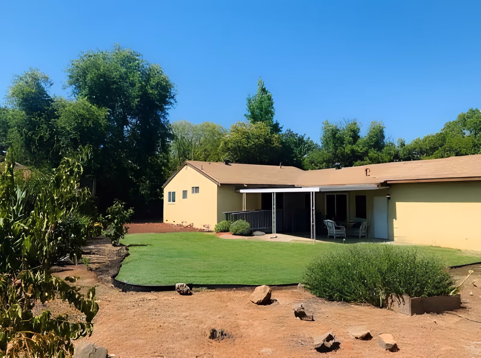 Single-story yellow building with a covered patio area furnished with chairs, surrounded by a grassy lawn and various bushes and trees under a clear blue sky.