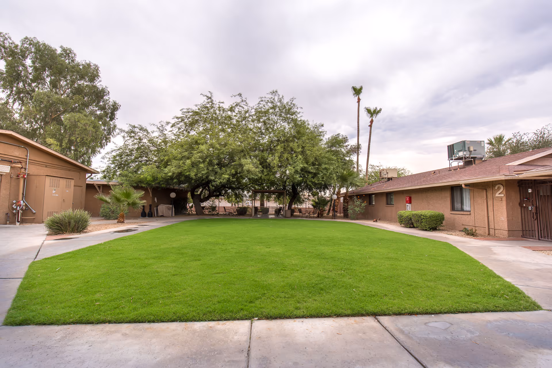 A courtyard area with a well-maintained green lawn surrounded by single-story buildings. There are trees providing shade in the background and a concrete walkway around the grass. The sky is cloudy.