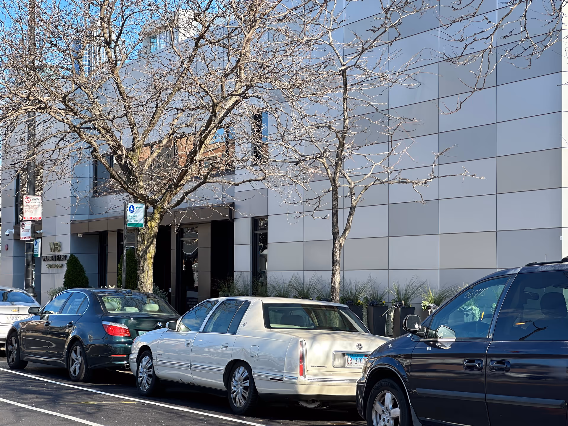 Front exterior of a modern building with leafless trees and several cars parked along the street.