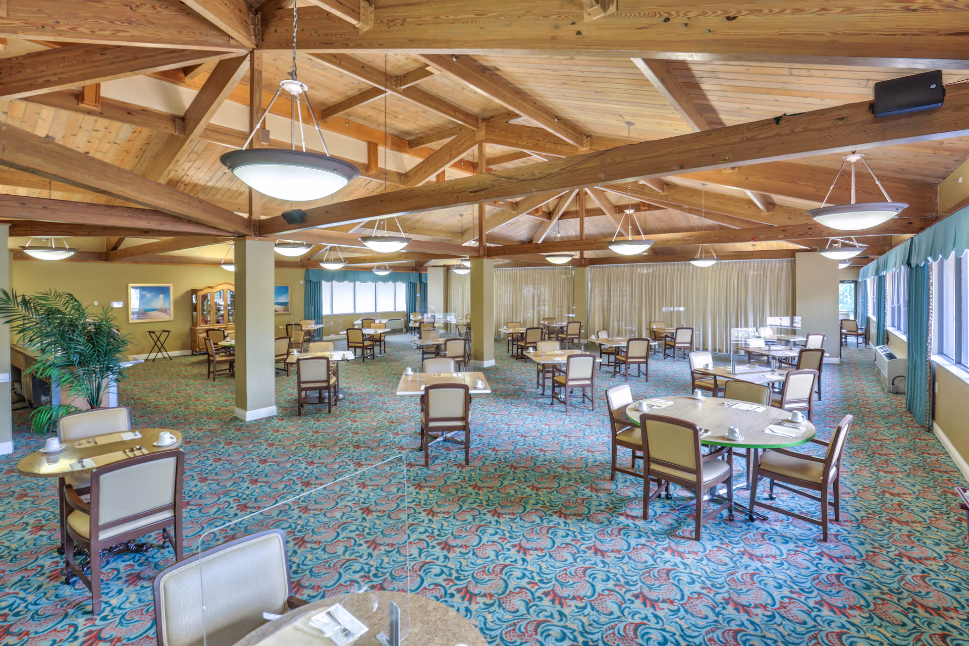 Spacious dining room with multiple tables and chairs arranged neatly on a patterned carpet. The room features a high wooden ceiling with exposed beams and several hanging light fixtures. Large windows with teal curtains allow natural light to fill the space, and there are plants and framed pictures on the walls.