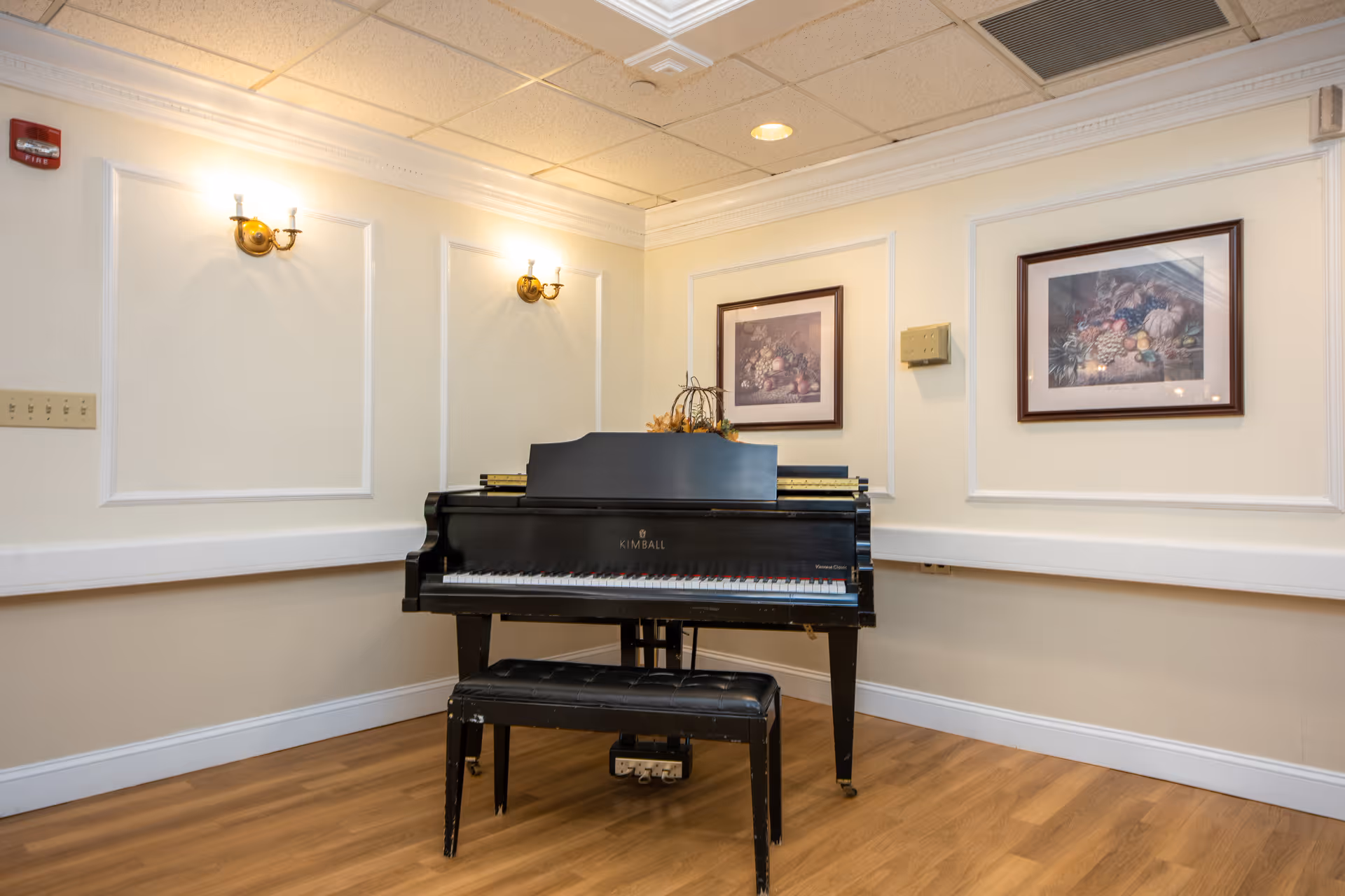 A black Kimball piano with a matching bench is placed against a cream-colored wall in a room with wooden flooring. The wall features two framed paintings of fruit and floral arrangements, and two brass wall sconces with lights are mounted above the piano. The ceiling has a grid pattern with recessed lighting.