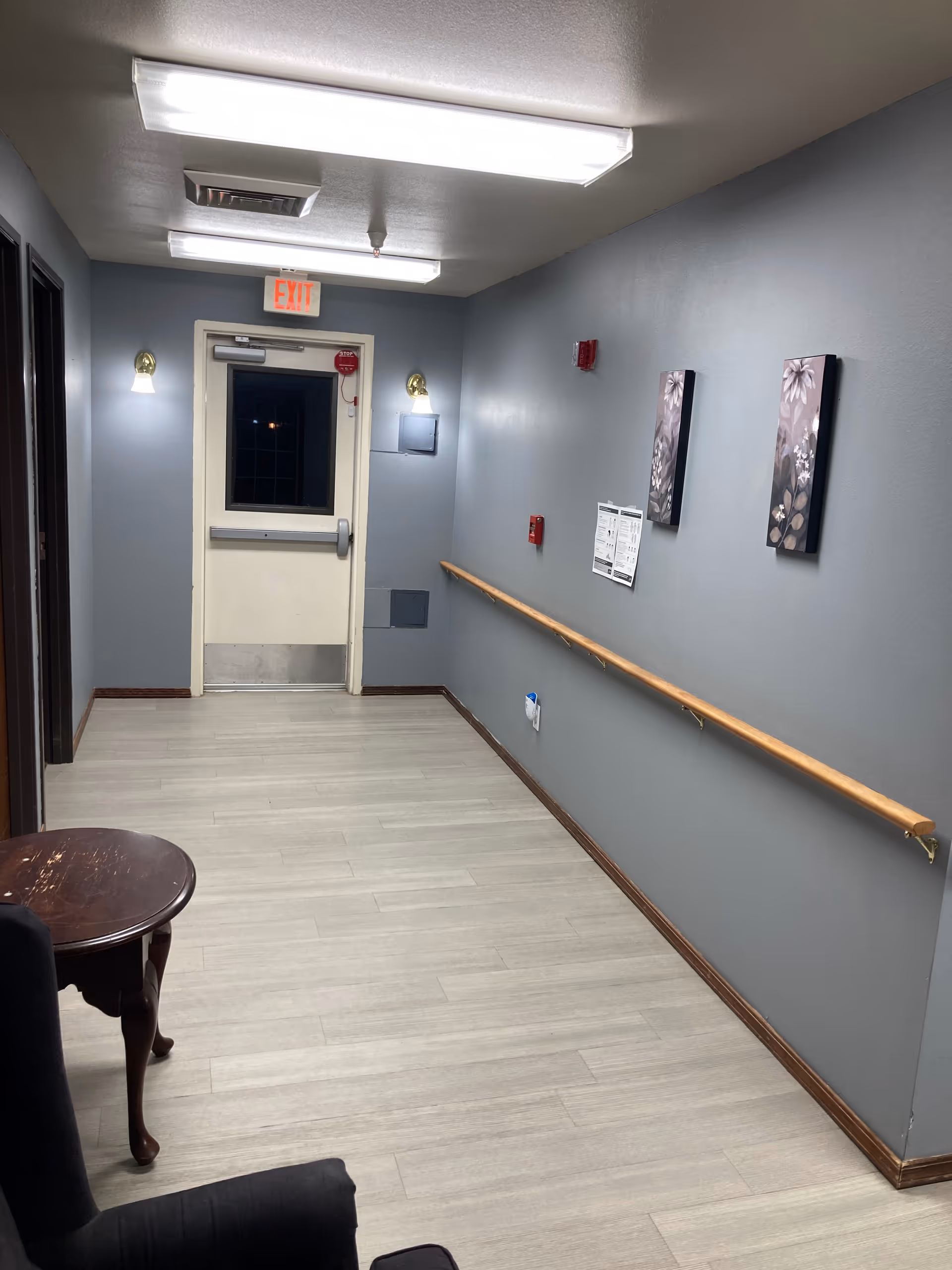 A hallway with light gray walls and light wood flooring. There is a wooden handrail along the right wall, two floral paintings, a fire alarm, and a posted notice. The hallway ends with a white door featuring a window and an illuminated exit sign above it. Two wall sconces provide additional lighting, and there is a small round wooden table and a dark upholstered chair partially visible in the foreground.