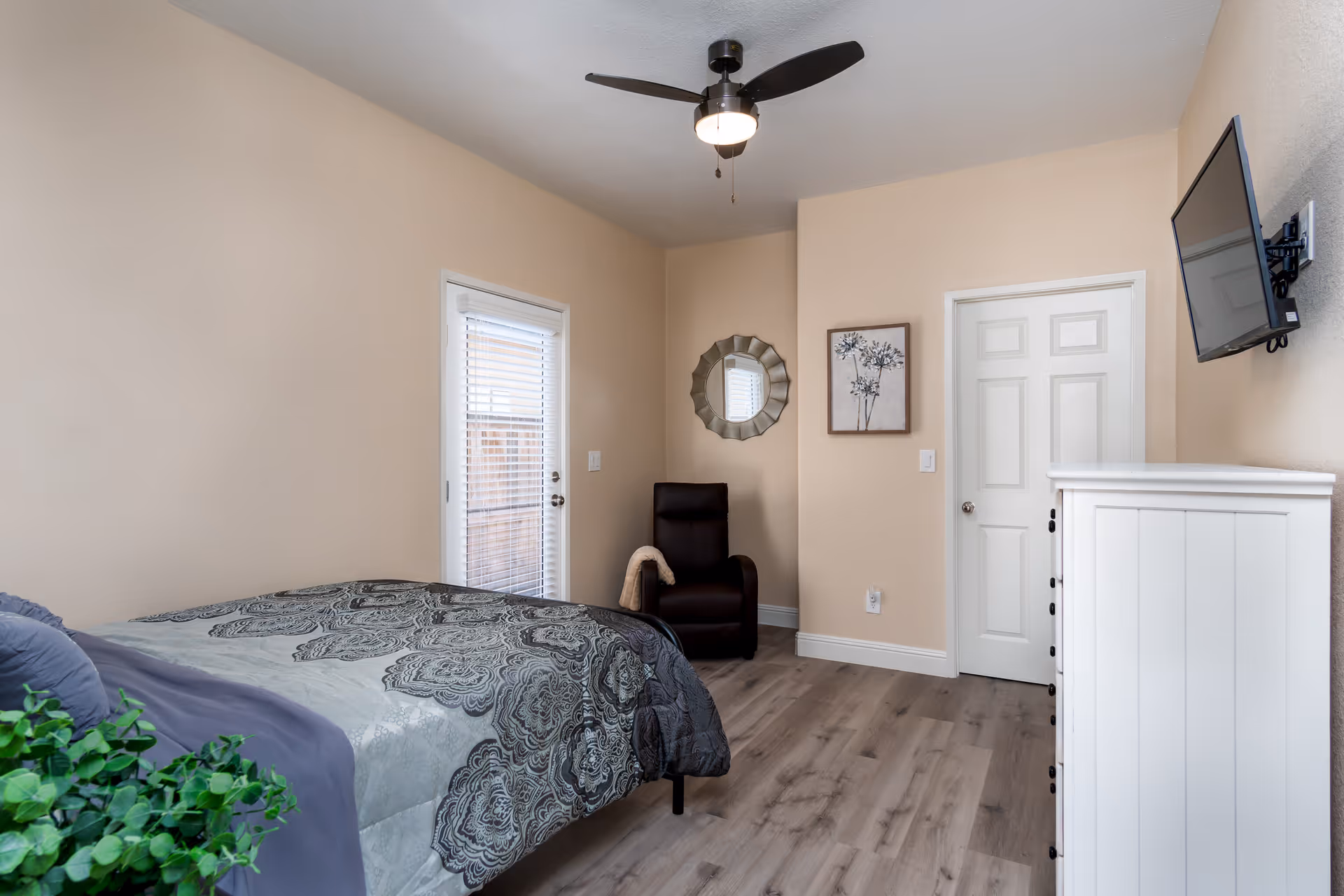 A bedroom with beige walls and wood flooring featuring a bed with patterned bedding and a green plant on the side. There is a black recliner chair in the corner next to a door with blinds, a decorative round mirror, and a framed floral picture on the wall. A white dresser is positioned under a wall-mounted flat-screen TV.