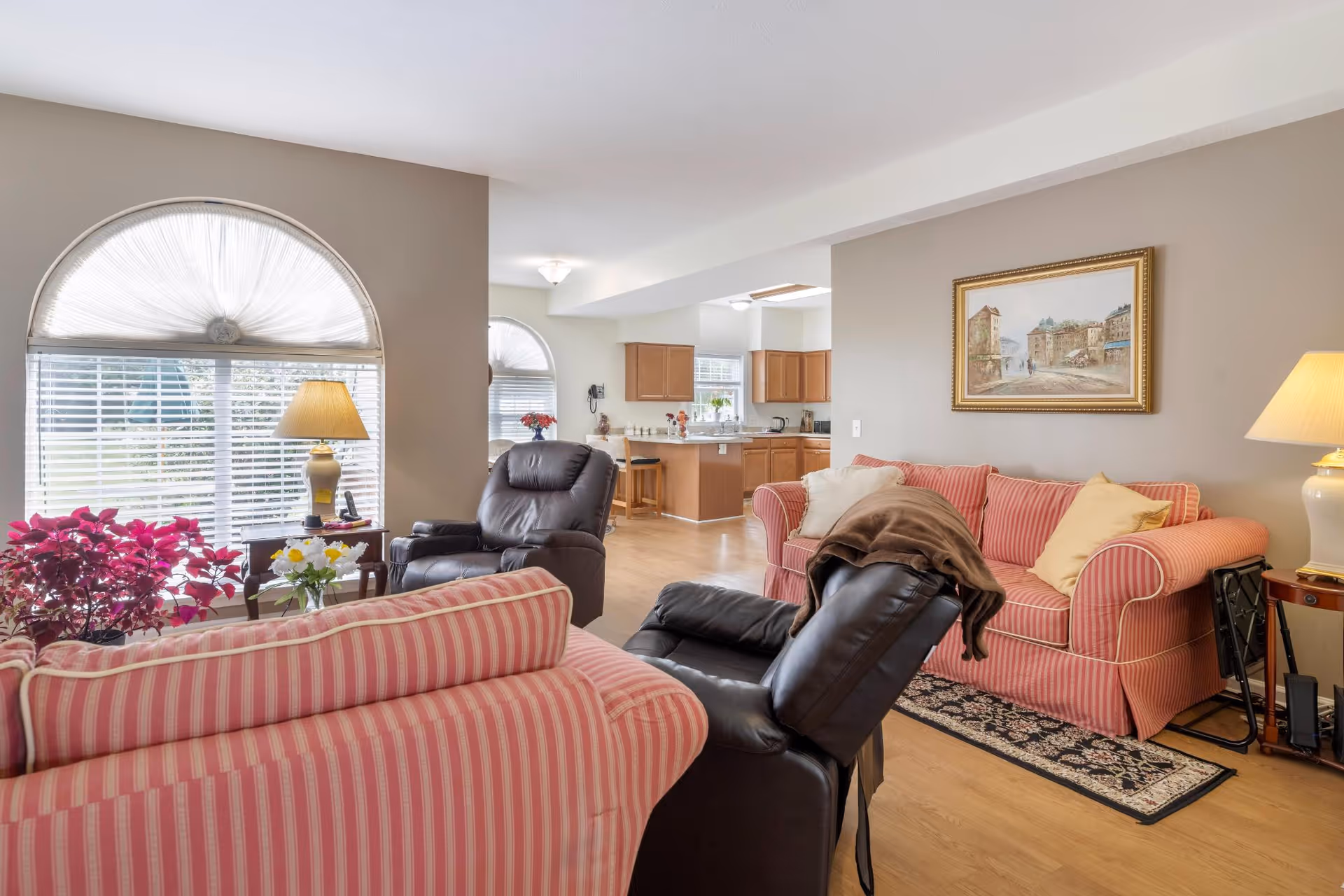 Sunlit living room with pink striped sofas, a black leather recliner, lamps, houseplants and an open view into the kitchen.