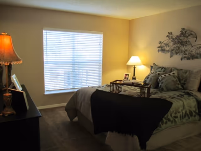 A cozy bedroom with a large window covered by white blinds. The bed is made with green and black patterned bedding and multiple pillows. A wooden tray with a book and a photo frame sits on the bed. There is a bedside table with a lamp and a decorative metal wall art above the bed. A dresser with a lamp and picture frames is visible on the left side of the room.