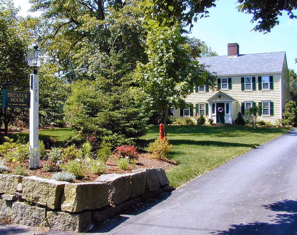 A two-story yellow house with green shutters and a chimney, surrounded by a well-maintained lawn and various trees and shrubs. A paved driveway leads up to the house. A white post with a lantern and a sign reading 'Decatur House 176 Main Street' is visible near the driveway entrance.