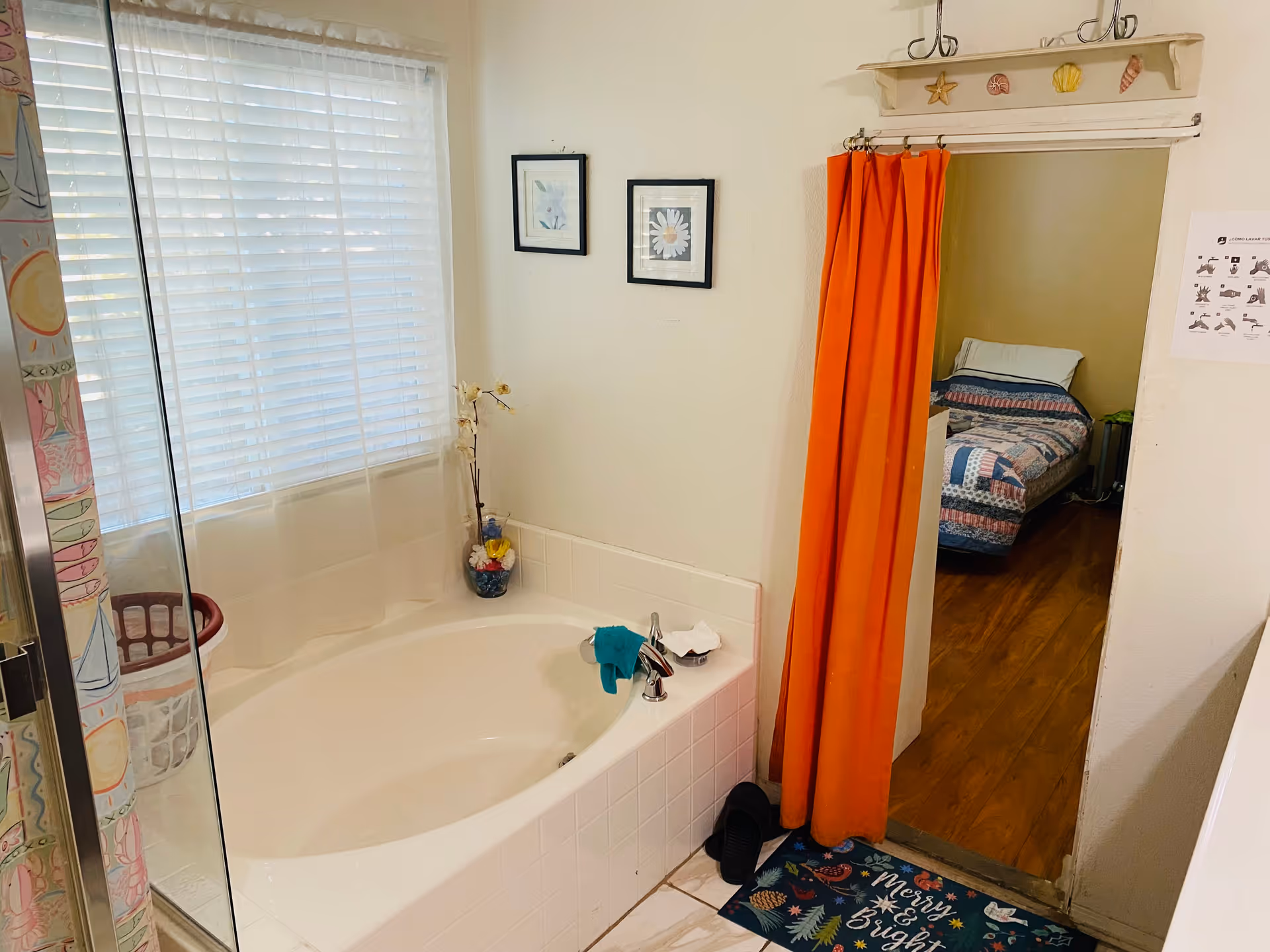 A bathroom with a white bathtub next to a window with white blinds and a sheer curtain. There is a vase with artificial flowers on the bathtub ledge. Two framed floral pictures hang on the wall above the bathtub. An orange curtain hangs in the doorway leading to a bedroom with a bed covered in a patchwork quilt. A colorful floor mat with the words 'Merry & Bright' is placed near the doorway.