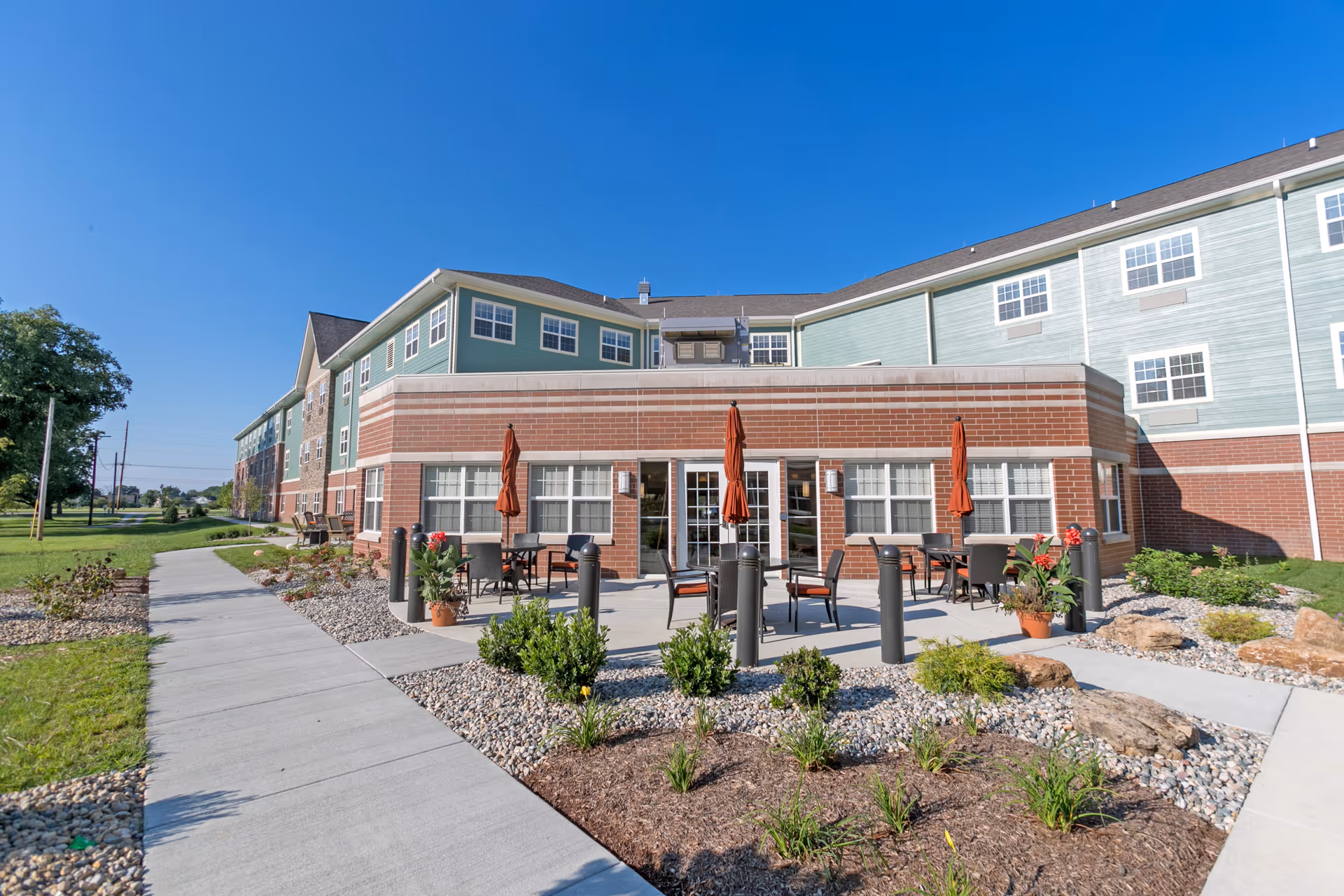 Exterior front view of a senior living building with a patio area featuring tables, umbrellas and landscaping along a sidewalk.