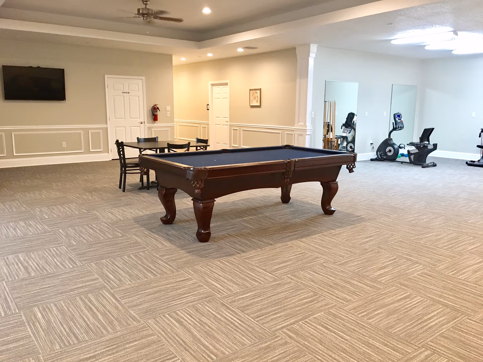 A spacious recreational room with a pool table in the center, a small table with chairs against the wall, a wall-mounted TV, and exercise equipment including stationary bikes near two large mirrors. The room has beige walls, carpeted floor with a patterned design, and ceiling fans.