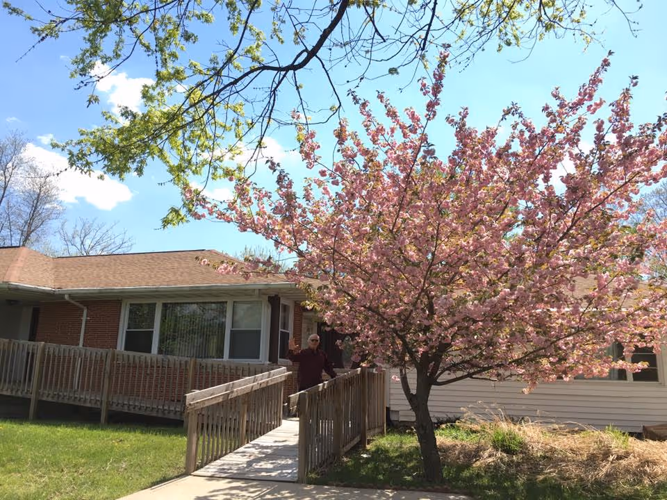 A single-story brick and siding building with a wooden ramp leading to the entrance. A person is standing on the ramp waving. In front of the building, there is a tree with pink blossoms and green grass surrounding the area under a clear blue sky.