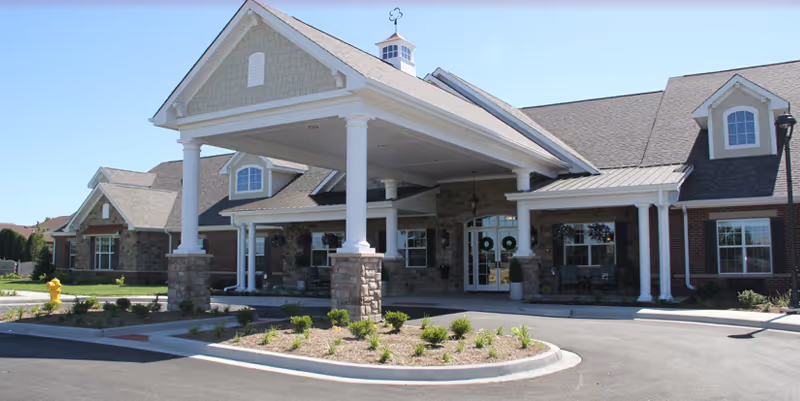 Front exterior view of Bickford of Spotsylvania facility showing a large covered entrance supported by white columns, stone and brick facade, multiple windows, and a landscaped area with small plants and a fire hydrant near the driveway.