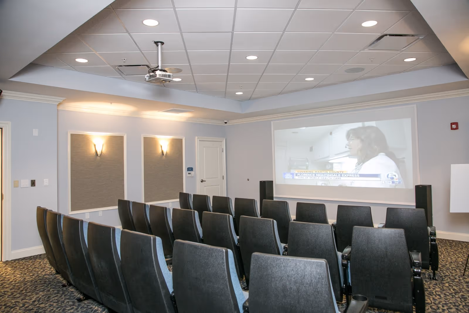 A small theater room with multiple rows of black cushioned seats facing a large screen on the wall. The screen displays a news segment with a woman speaking. The room has a drop ceiling with recessed lighting and a ceiling-mounted projector.