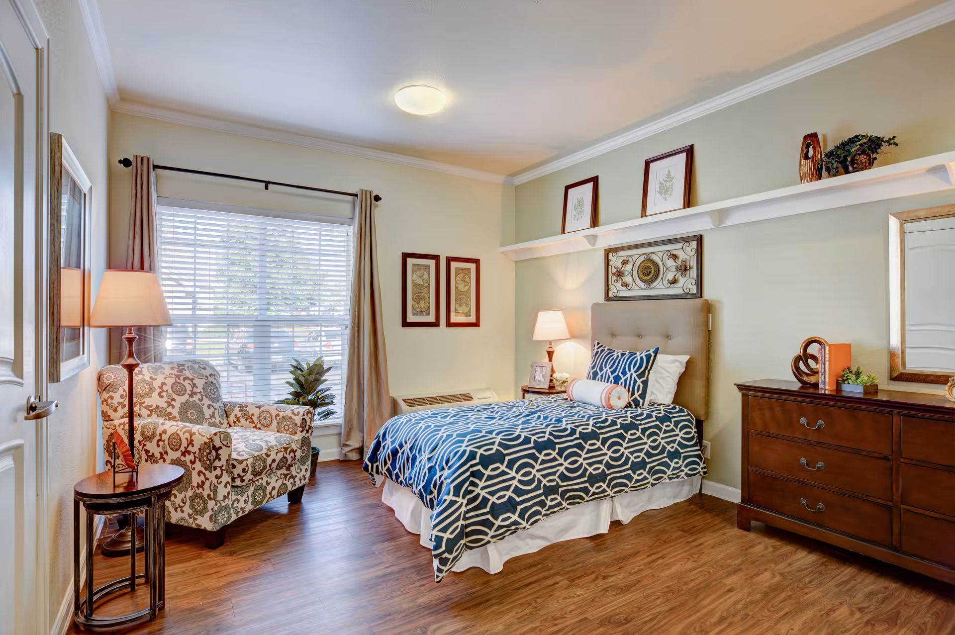 Sunny furnished bedroom with a patterned armchair, single bed with blue geometric bedding, dresser, and a large window with curtains.