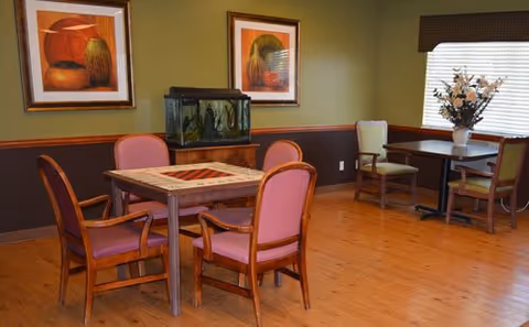 A cozy room with wooden flooring featuring two tables with chairs. One table has four pink cushioned chairs around it, and the other table near the window has two green cushioned chairs. There is a fish tank on a wooden cabinet against the wall, two framed paintings above it, and a vase with flowers on the table by the window with blinds partially open.