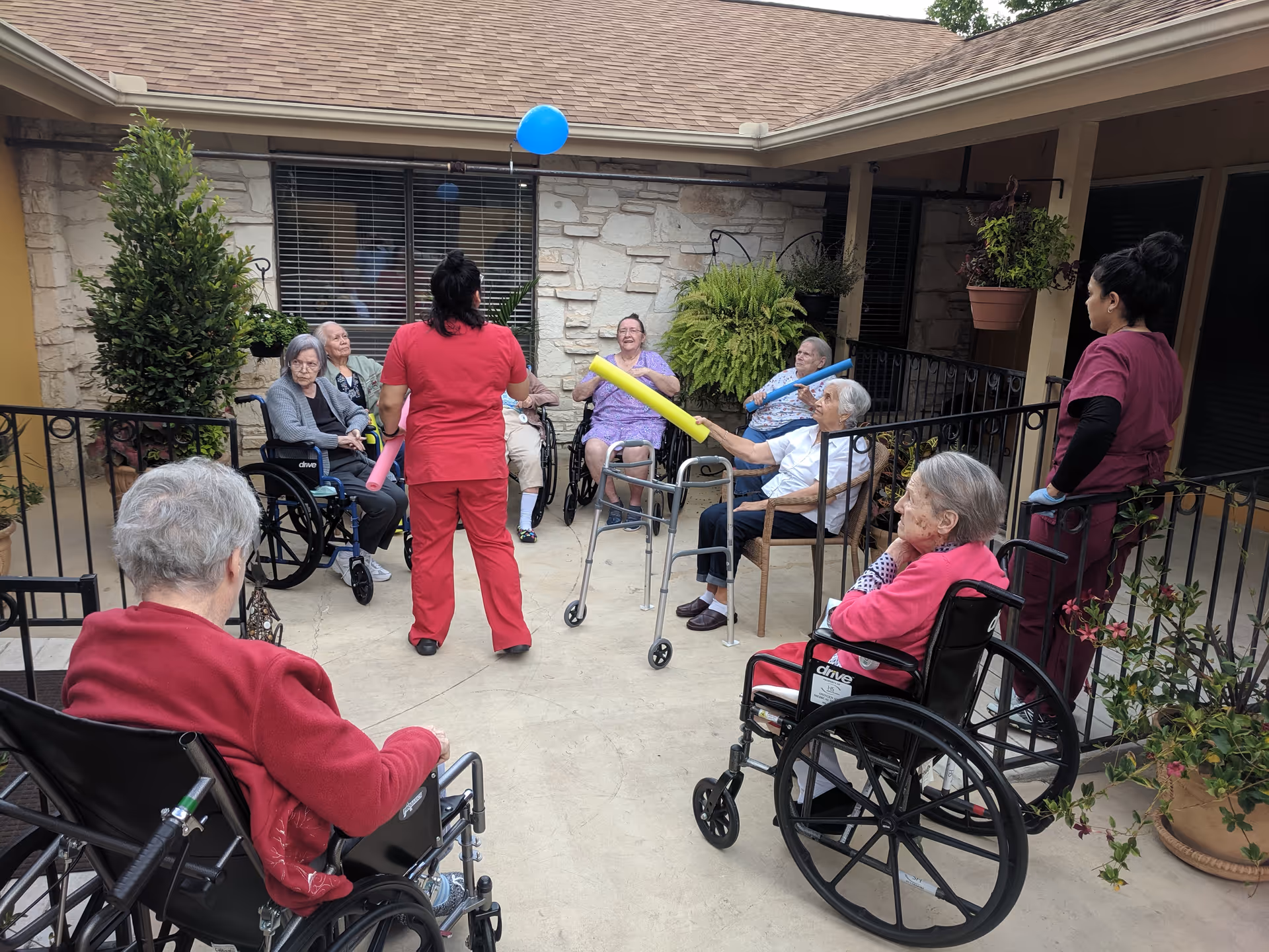 Seniors in wheelchairs and staff in scrubs play with a balloon and foam noodles in an outdoor courtyard.