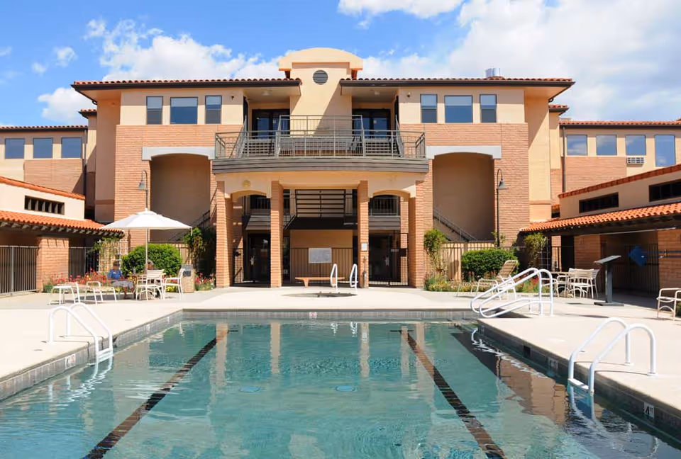 Outdoor swimming pool with clear water in front of a multi-story assisted living facility building under a partly cloudy sky. There are poolside chairs, tables with umbrellas, and some greenery around the pool area.