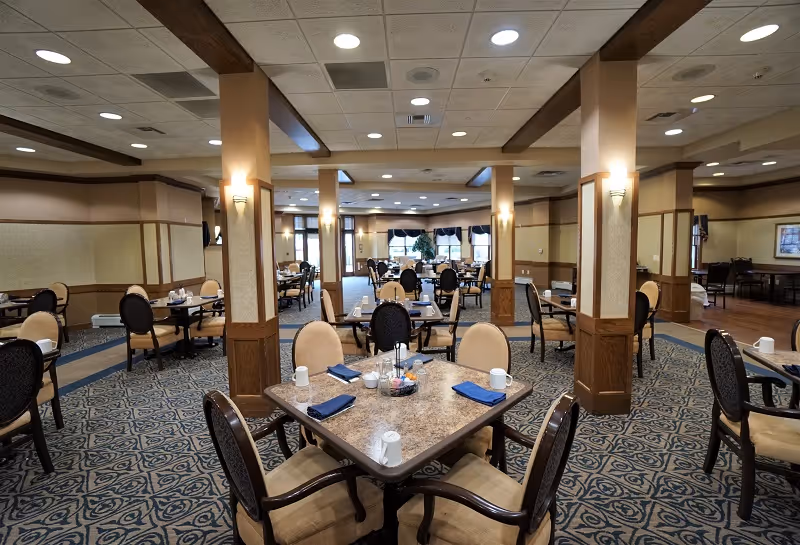 A spacious dining room with multiple square tables set with blue napkins, white mugs, and condiment holders. The room features patterned carpet, beige walls with wooden paneling, and several columns. Large windows with dark valances allow natural light to enter the room.
