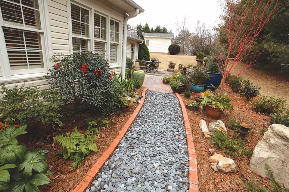 A garden pathway lined with small red bricks and filled with gray gravel leads to a patio area with outdoor furniture. The path is bordered by various plants, shrubs, and potted flowers on both sides. The side of a light-colored building with multiple windows is visible on the left, and trees and grass are seen in the background.
