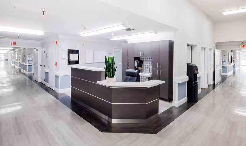 A bright and clean nursing facility hallway with a central reception desk featuring a potted plant. The hallway extends in two directions with multiple doors, exit signs, and a water dispenser visible. The walls are painted white with a light blue lower section, and the floor has a light wood finish with a darker wood section around the reception area.