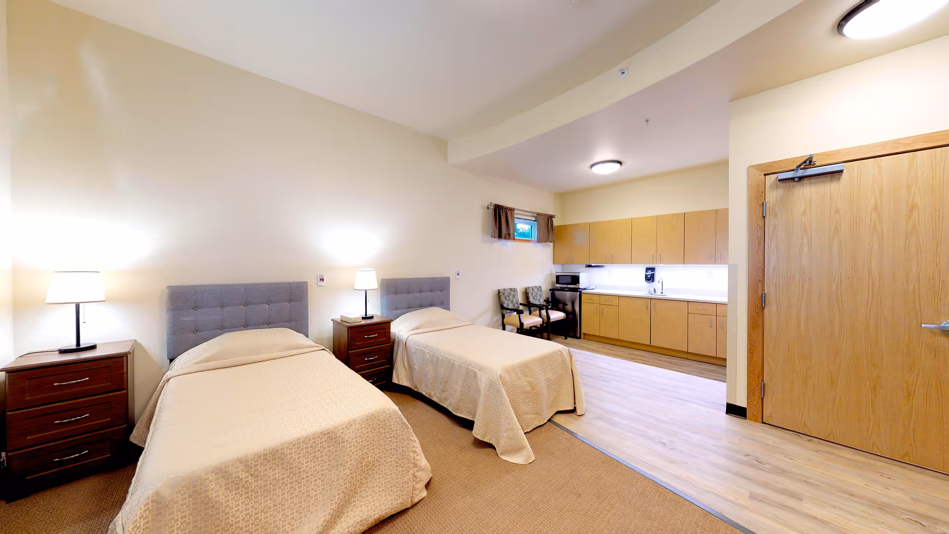 A well-lit assisted living room featuring two single beds with beige bedspreads and gray tufted headboards. Each bed has a wooden nightstand with a lamp. The room includes a small kitchenette with wooden cabinets, a microwave, a coffee maker, and two upholstered chairs near a small window with a brown valance. The floor transitions from carpet near the beds to wood laminate near the kitchenette and door.