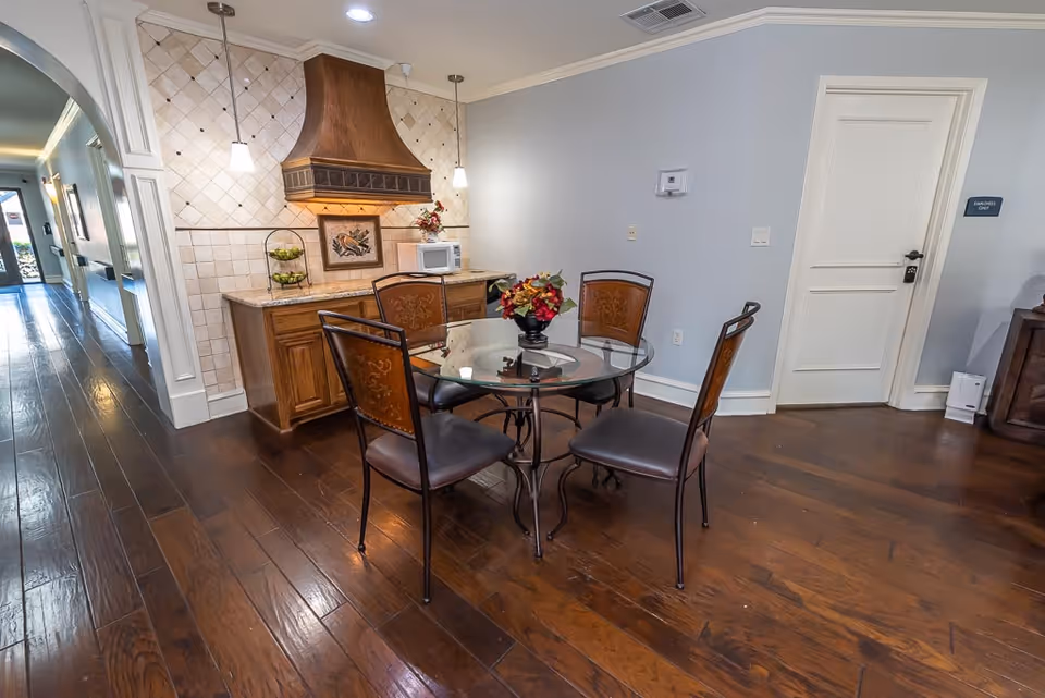 A small dining area with a round glass table and four metal chairs with brown cushioned seats. Behind the table is a wooden cabinet with a decorative range hood above it, a microwave, and a two-tier fruit basket. The walls are light gray with white trim, and the floor is dark hardwood. A hallway is visible to the left, and a closed white door with a sign reading 'Employees Only' is on the right.