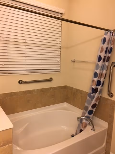 A bathroom featuring a white bathtub with beige tiled walls surrounding it. There is a metal grab bar on the wall above the tub and a shower curtain with a pattern of large colored circles in shades of blue, brown, and beige. A window with closed white blinds is above the grab bar, and a towel rack is mounted on the adjacent wall.