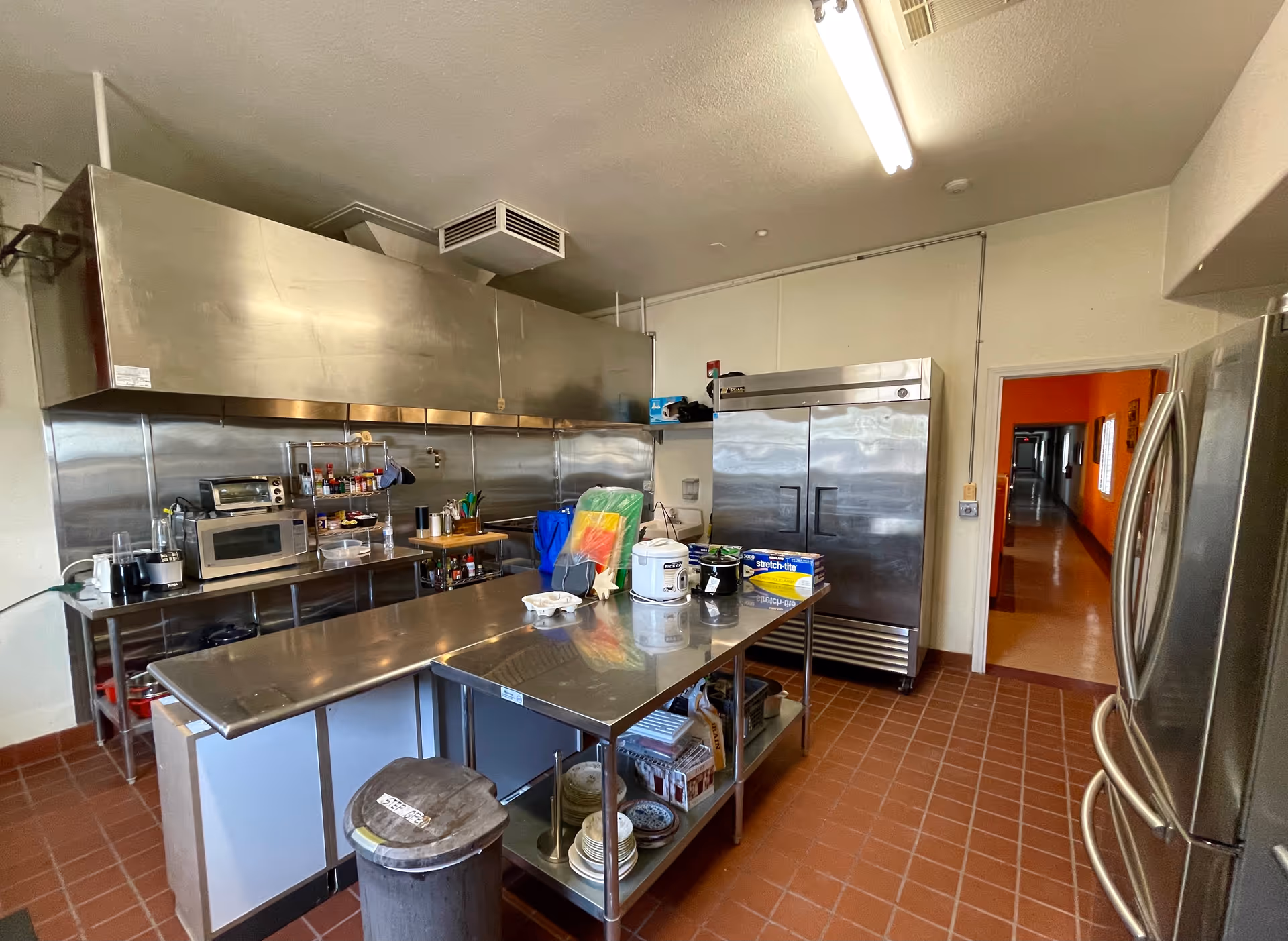 Interior view of a commercial kitchen with stainless steel appliances and countertops. There are two large refrigerators, a microwave, toaster oven, blender, and various kitchen utensils and supplies on the counters and shelves. The floor is tiled with reddish-brown tiles, and there is a hallway visible through a doorway.