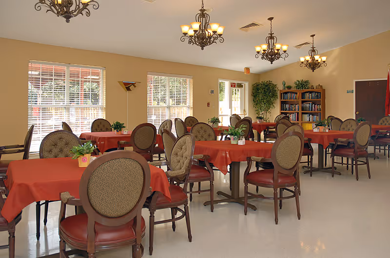 A well-lit dining room with multiple round tables covered in red tablecloths, each surrounded by cushioned chairs. The room has large windows with blinds, several chandeliers hanging from the ceiling, and a bookshelf filled with books against one wall. There are also some potted plants on the tables and in the corners of the room.