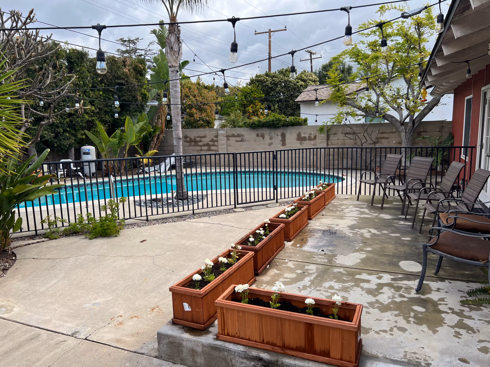 Outdoor patio area with planter boxes, chairs, string lights and a fenced swimming pool.